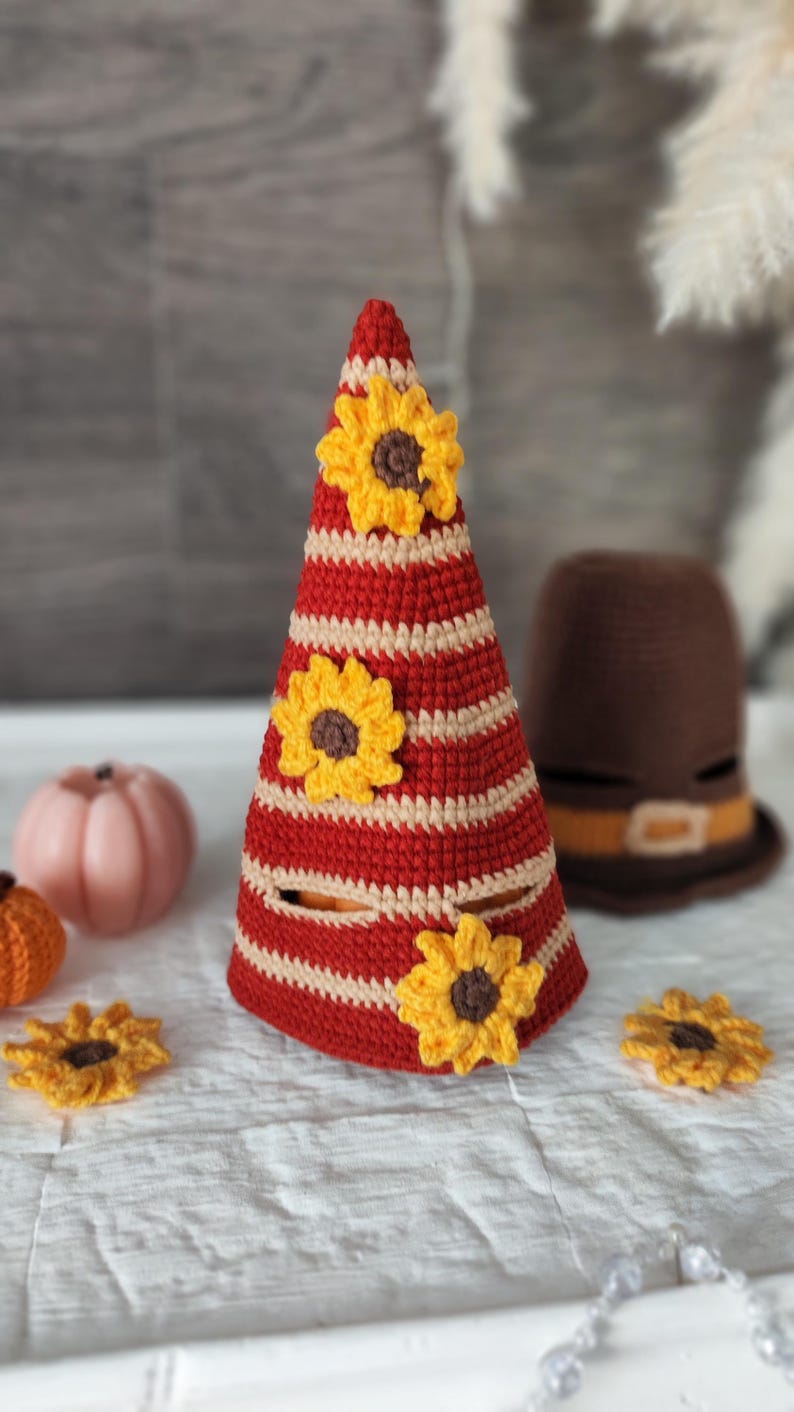 May include: A handmade crochet cone-shaped decoration with red and cream stripes, adorned with yellow and brown sunflowers. A brown crochet hat with a tan band is in the background. Other crochet pumpkins and sunflowers are also visible.