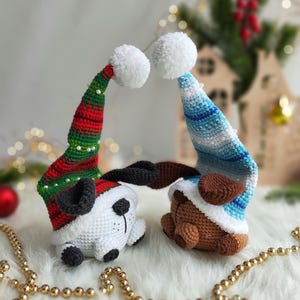 May include: Two crocheted stuffed animals wearing Santa hats. The animal on the left is white with black ears and a black nose. The animal on the right is brown with black ears and a black nose. Both animals are wearing red and green Santa hats with white pom-poms.