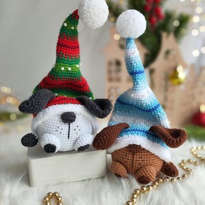 May include: Two crocheted dog ornaments wearing festive hats. One dog is white with black ears and a red and green hat with a white pom-pom. The other dog is brown with brown ears and a blue and white striped hat with a white pom-pom.