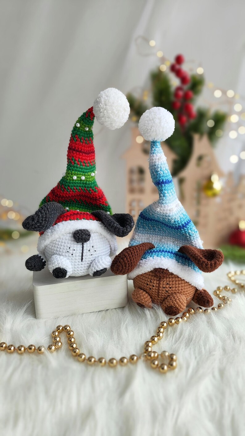 May include: Two crocheted dog ornaments wearing festive hats. One dog is white with black ears and a red and green hat with a white pom-pom. The other dog is brown with brown ears and a blue and white striped hat with a white pom-pom.