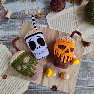 May include: Three crocheted Halloween-themed coffee cups with candy corn accents. The cups are decorated with different skull designs: a white skull with black eyes, an orange skull with brown dripping icing, and a green skull with brown dripping icing.