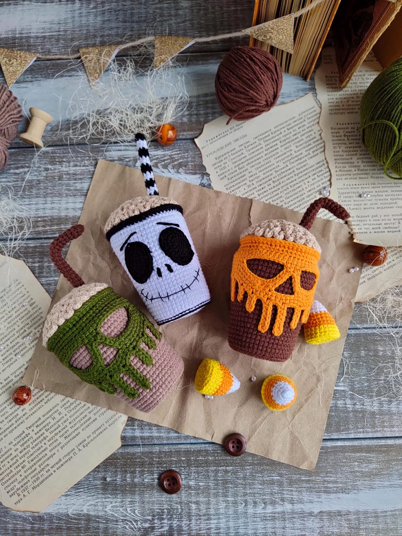 May include: Three crocheted Halloween-themed coffee cups with candy corn accents. The cups are decorated with different skull designs: a white skull with black eyes, an orange skull with brown dripping icing, and a green skull with brown dripping icing.