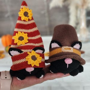 May include: Two handmade crochet cat gnomes. One gnome wears a red and tan striped hat with yellow sunflower accents. The other gnome wears a brown hat with a yellow and orange band. Both gnomes are black with pink noses and ears.