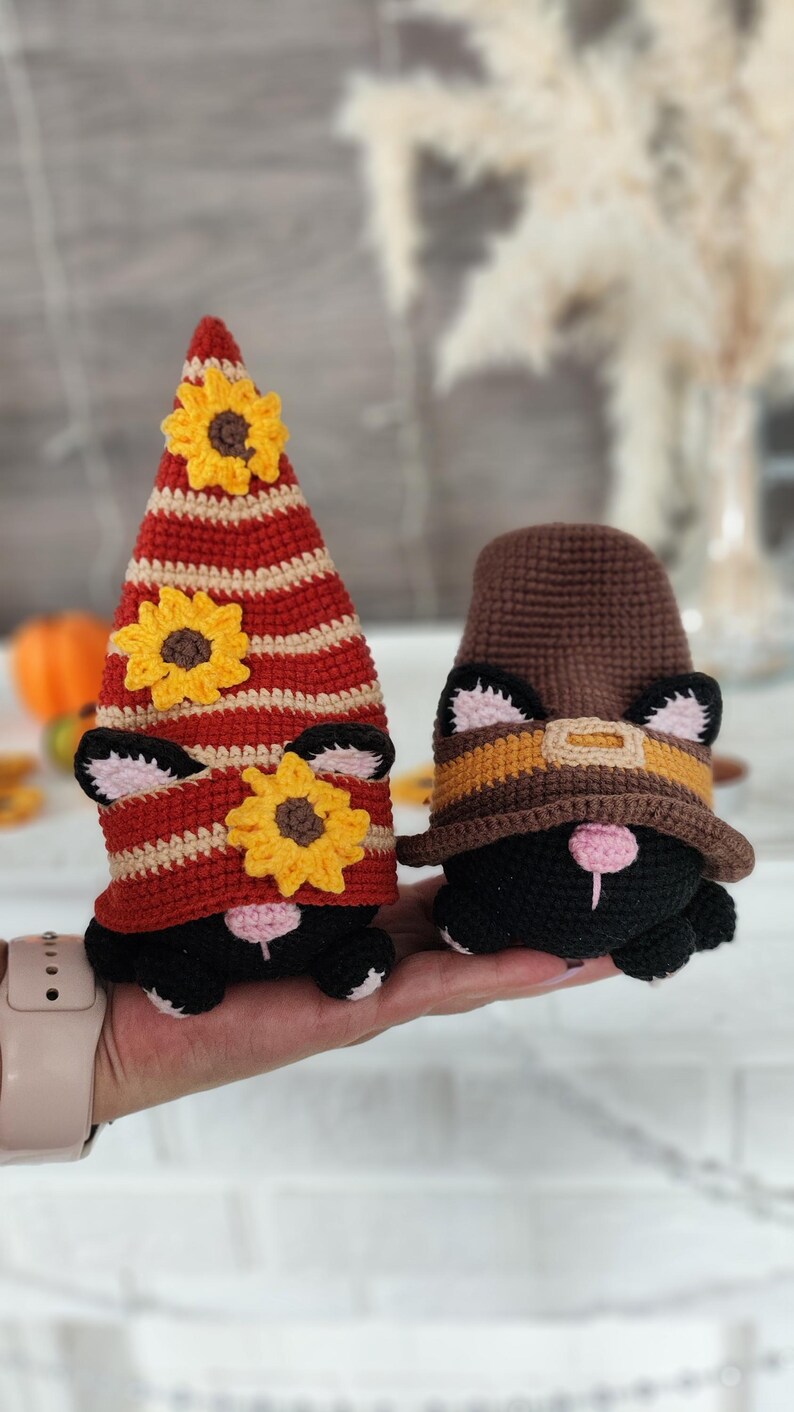 May include: Two handmade crochet cat gnomes. One gnome wears a red and tan striped hat with yellow sunflower accents. The other gnome wears a brown hat with a yellow and orange band. Both gnomes are black with pink noses and ears.