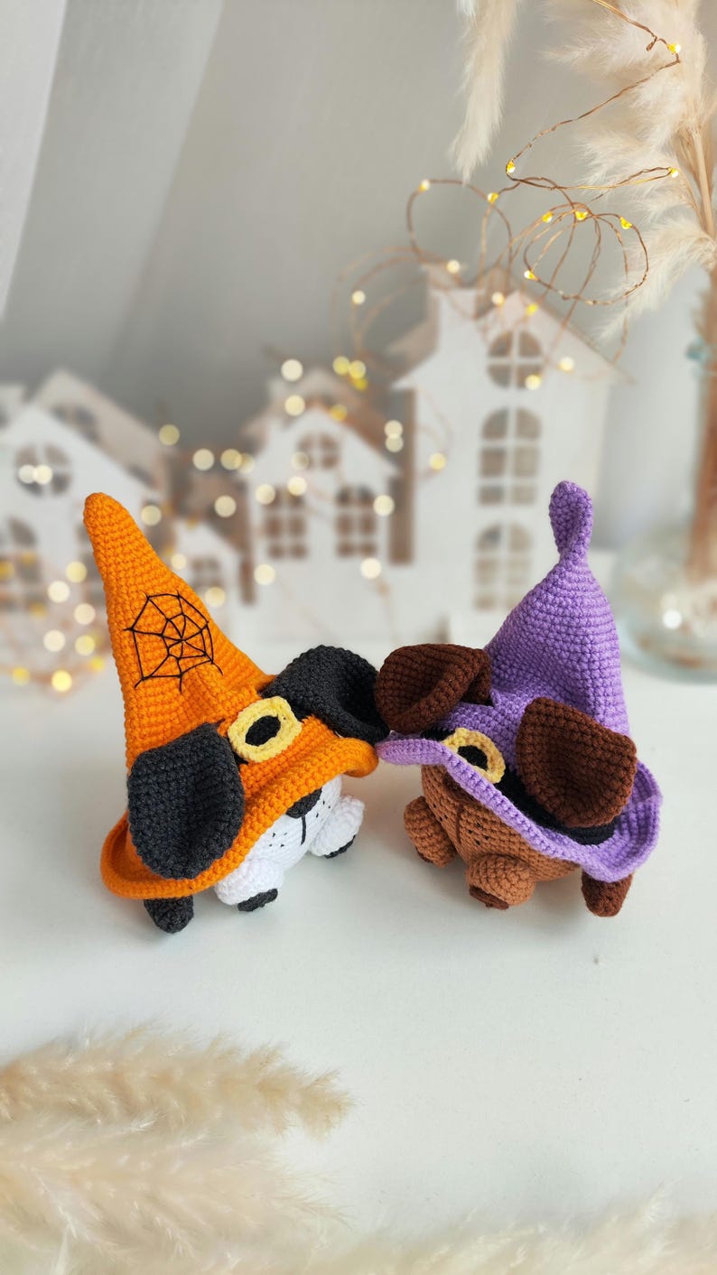 May include: Two crocheted Halloween dog toys wearing witch hats. One hat is orange with a black spiderweb and the other is purple. Both dogs have brown ears and black noses.