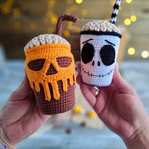 May include: Two crocheted Halloween-themed coffee cups. One cup is decorated with a pumpkin face and the other with a skull face. Both cups have a crocheted straw.
