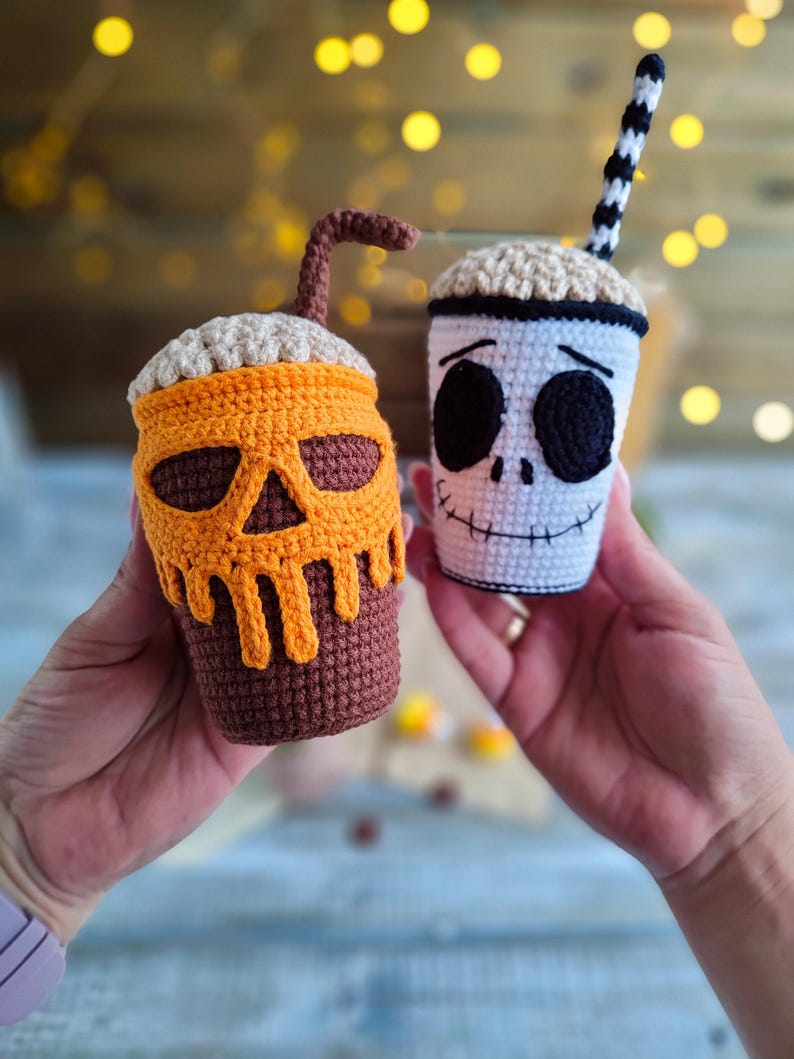 May include: Two crocheted Halloween-themed coffee cups. One cup is decorated with a pumpkin face and the other with a skull face. Both cups have a crocheted straw.