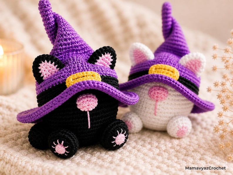 May include: Two handmade crochet cat figures, one black and one white, wearing purple witch hats with yellow bands. The cats have pink ears and noses, and are sitting on a soft, textured surface. The figures are whimsical and decorative.