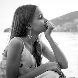 May include: A young woman with long hair is wearing a white tank top with a small embroidered design. She is wearing a silver triangle earring and a silver bracelet. She is looking off to the side and has a thoughtful expression on her face.