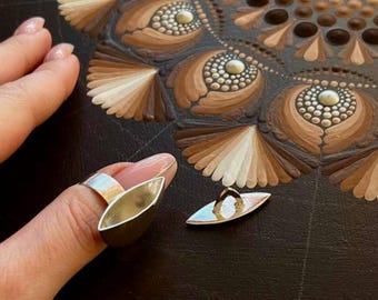 Puede incluir: Un anillo de plata con un diseño en forma de barco. El anillo está en un dedo y el barco está abierto. Hay un colgante de plata a juego en la mesa.
