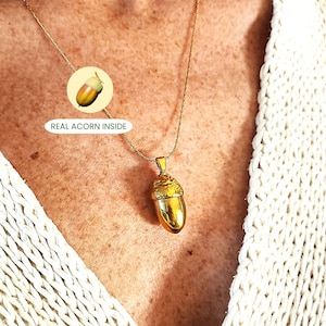 May include: A gold acorn pendant necklace. The pendant, a detailed replica of an acorn, has a smooth, polished body and a textured cap. The necklace is worn on a delicate gold chain. Text on the image reads: REAL ACORN INSIDE.