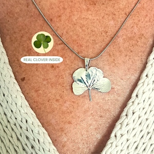 May include: A silver-toned necklace featuring a pendant shaped like a three-leaf clover. The pendant has a metallic finish and is suspended from a thin chain. A small circular graphic above the pendant shows a green clover with the text REAL CLOVER INSIDE.