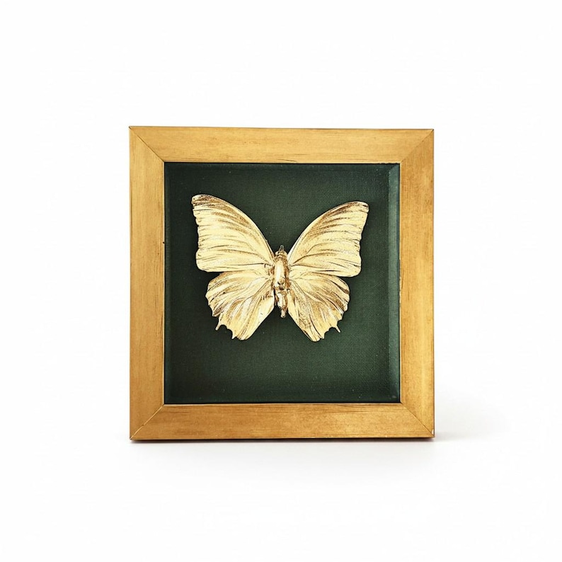 May include: A square wall art piece featuring a three-dimensional gold-coloured butterfly mounted against a dark green fabric background, enclosed within a simple gold-toned wooden frame with clean lines and a smooth finish.