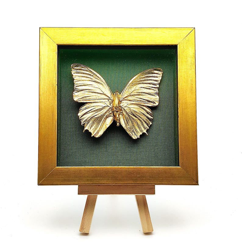 May include: A gold-coloured butterfly sculpture mounted inside a square frame with a metallic gold finish and a dark green fabric backing. The framed butterfly is displayed on a small, light-coloured wooden tripod easel stand against a white background.
