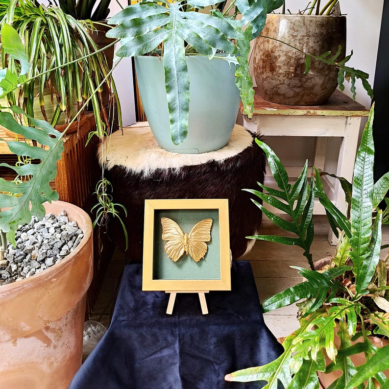 May include: A square wooden frame with a light tan finish containing a gold-coloured butterfly sculpture mounted against a dark green background. The framed artwork is displayed on a small wooden easel stand, positioned upright for viewing.