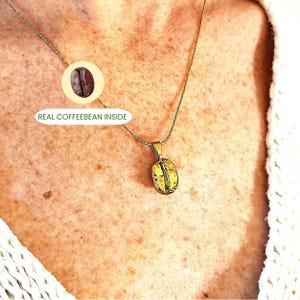 May include: A gold-colored pendant necklace featuring a coffee bean design. The pendant hangs from a delicate silver chain. The image includes the text "REAL COFFEEBEAN INSIDE" next to a graphic of a coffee bean.