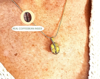 May include: A gold-colored pendant necklace featuring a coffee bean design. The pendant hangs from a delicate silver chain. The image includes the text "REAL COFFEEBEAN INSIDE" next to a graphic of a coffee bean.