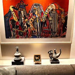 May include: A framed painting featuring three figures in colourful attire with the text AND AFRICAN TEA, displayed above a shelf holding a grey lidded vessel with a small figure, a small framed butterfly specimen, and a metallic horse sculpture.