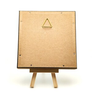 May include: A square frame backing made of light brown cardboard with a triangular gold metal hanging hook centred at the top. The frame is supported by a small, light-coloured wooden easel stand, showing the back side of the display object.