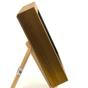 May include: A rectangular box with a metallic gold finish on the exterior sides, positioned at an angle and supported by a light-coloured wooden easel stand. The interior of the box is dark, and the structure is shown from a side profile view.