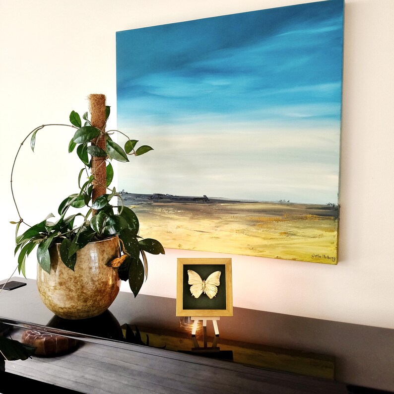 May include: A square landscape painting featuring a bright blue sky above a golden yellow field, displayed alongside a potted green plant in a textured brown pot and a small framed white butterfly specimen on a miniature wooden easel.