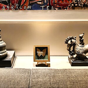 May include: A gold-coloured butterfly specimen displayed inside a square wooden frame with a dark green backing, resting on a small wooden easel. The frame is positioned between a dark metal bell-shaped object and a bronze-toned horse sculpture.
