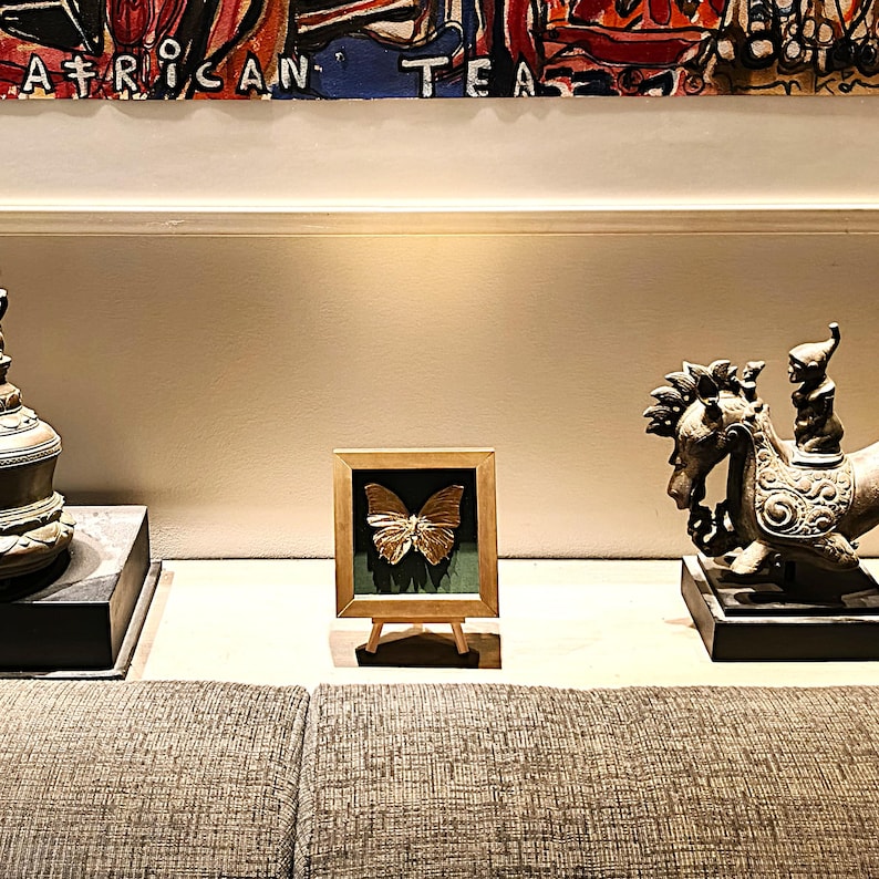 May include: A gold-coloured butterfly specimen displayed inside a square wooden frame with a dark green backing, resting on a small wooden easel. The frame is positioned between a dark metal bell-shaped object and a bronze-toned horse sculpture.