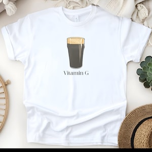Może przedstawiać: Biała koszulka z grafiką ciemnego piwa stout w szklance typu pint z pianą. Tekst "Vitamin G" jest wydrukowany pod ilustracją piwa. Koszulka jest wykonana z miękkiego materiału.