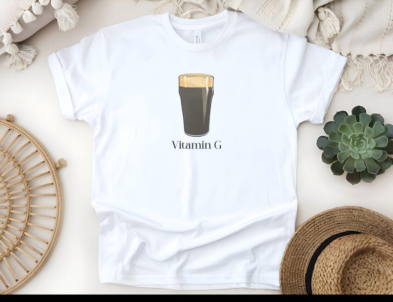 Może przedstawiać: Biała koszulka z grafiką ciemnego piwa stout w szklance typu pint z pianą. Tekst "Vitamin G" jest wydrukowany pod ilustracją piwa. Koszulka jest wykonana z miękkiego materiału.