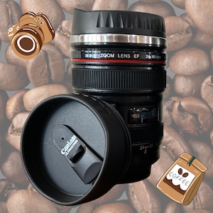 May include: Black camera lens shaped travel mug with a black lid and a silver band around the top. The mug has the text "ZOOM LENS EF 24-105mm" printed on it. The mug is sitting on a background of coffee beans.