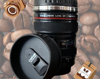 May include: Black camera lens shaped travel mug with a black lid and a silver band around the top. The mug has the text "ZOOM LENS EF 24-105mm" printed on it. The mug is sitting on a background of coffee beans.