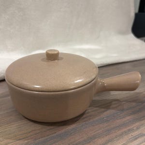 May include: A beige ceramic soup bowl with a lid and a handle. The bowl has a round shape and a small knob on top of the lid. The handle is attached to the side of the bowl. The bowl is sitting on a wooden surface.