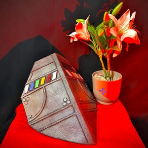 May include: A metallic grey, geometric sculpture with colourful rectangular accents. The sculpture is set on a red surface, with a potted plant featuring white and red flowers in the background. The overall aesthetic is modern and artistic.