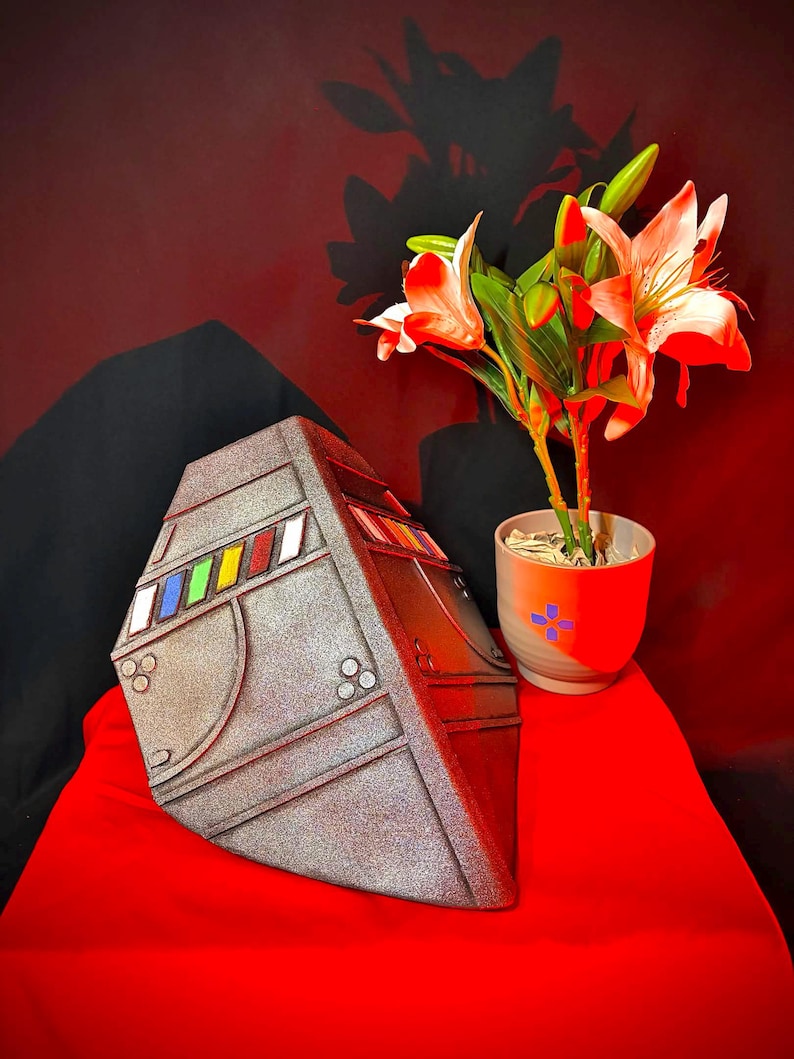 May include: A metallic grey, geometric sculpture with colourful rectangular accents. The sculpture is set on a red surface, with a potted plant featuring white and red flowers in the background. The overall aesthetic is modern and artistic.