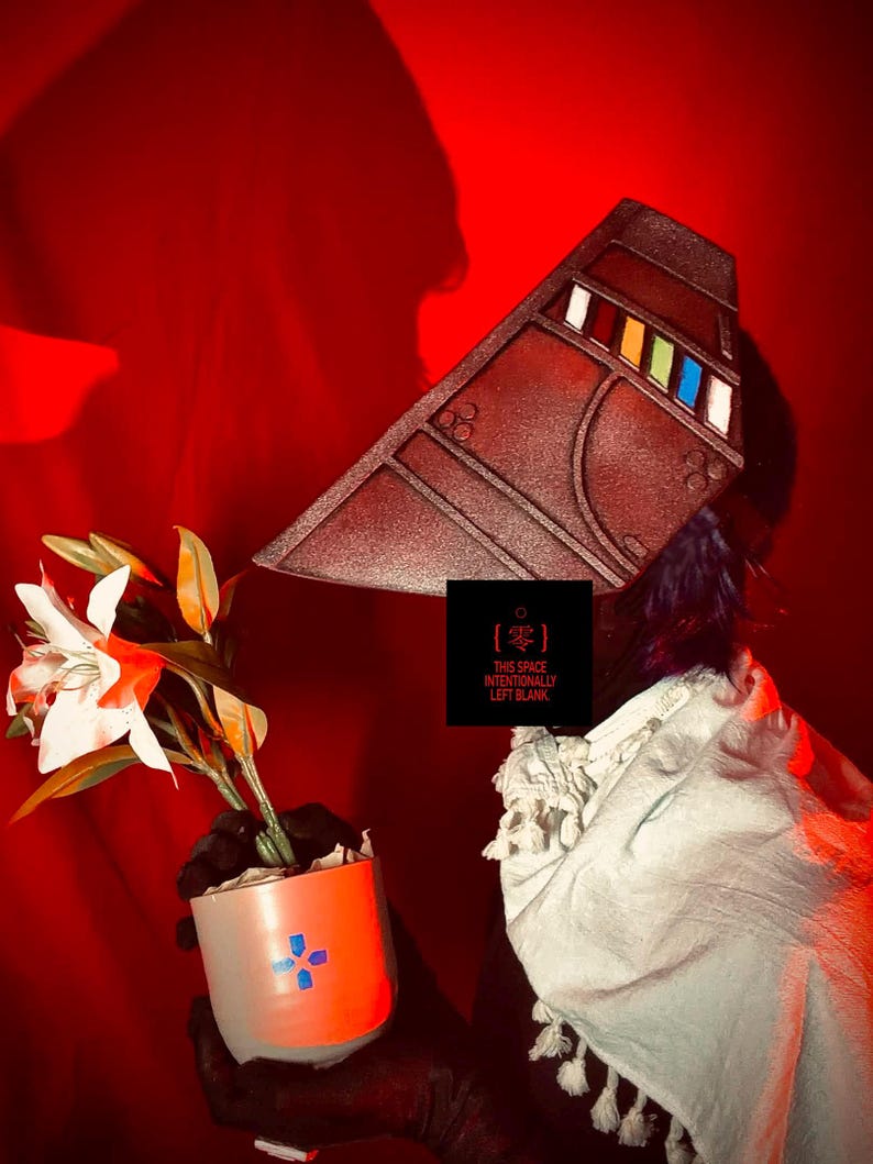 May include: A brown, angular headpiece with colourful accents and a white scarf. A small orange and white pot with a blue cross design holds a plant with white and orange flowers. The text "THIS SPACE INTENTIONALLY LEFT BLANK" is visible.