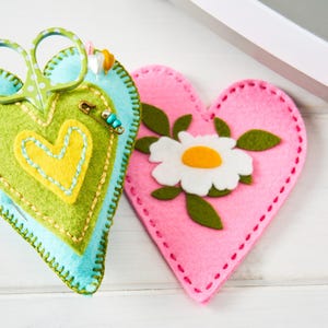 Peut inclure: Deux coussins &agrave; &eacute;pingles en forme de c&oelig;ur. L'un est bleu, vert et jaune avec des ciseaux et une &eacute;pingle de s&ucirc;ret&eacute;. L'autre est rose avec une marguerite blanche et des feuilles vertes. Les deux sont en feutre et cousus.
