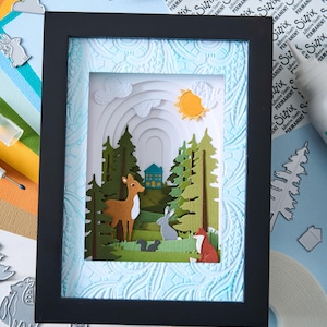K&ouml;nnte beinhalten: Eine gerahmte Papierkunstszene zeigt einen Wald mit einem Hirsch, einem Kaninchen und einem Fuchs. Das Kunstwerk zeigt geschichtete Papierauschnitte von B&auml;umen, einem Haus und einer Sonne. Der Rahmen ist schwarz und der Hintergrund hellblau mit einem strukturierten Muster.