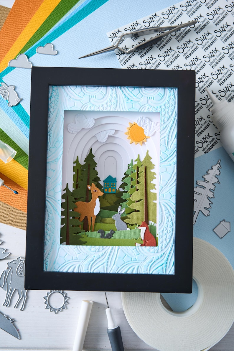 K&ouml;nnte beinhalten: Eine gerahmte Papierkunstszene zeigt einen Wald mit einem Hirsch, einem Kaninchen und einem Fuchs. Das Kunstwerk zeigt geschichtete Papierauschnitte von B&auml;umen, einem Haus und einer Sonne. Der Rahmen ist schwarz und der Hintergrund hellblau mit einem strukturierten Muster.