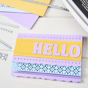 May include: A handmade greeting card with the word "HELLO" in lavender letters on a yellow and lavender background. The card has decorative edges and layers, with die-cut floral and stitched details. Craft supplies and a die-cutting machine are visible.