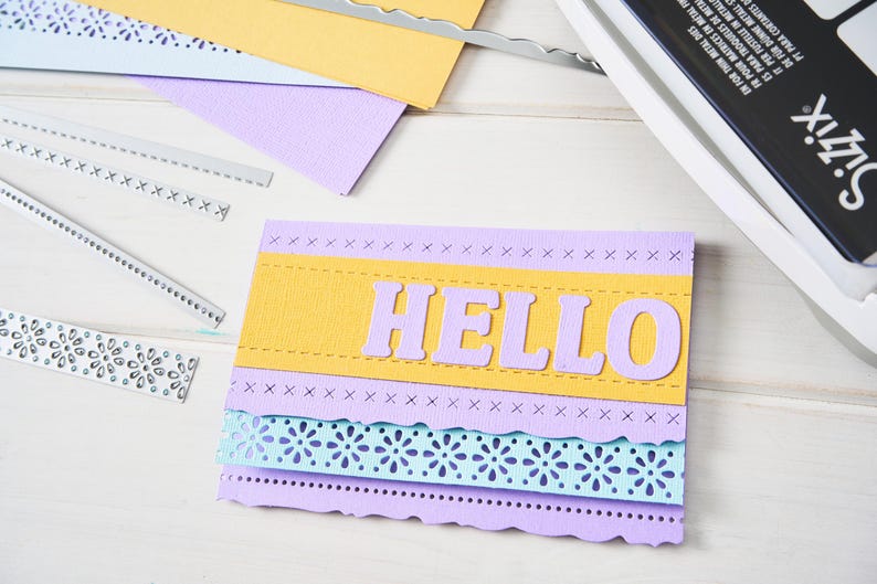 May include: A handmade greeting card with the word "HELLO" in lavender letters on a yellow and lavender background. The card has decorative edges and layers, with die-cut floral and stitched details. Craft supplies and a die-cutting machine are visible.