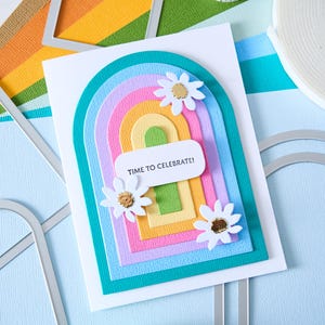 K&ouml;nnte beinhalten: Eine handgefertigte Gru&szlig;karte mit einem Regenbogenbogen-Design, mit Schichten aus farbigem Papier und wei&szlig;en G&auml;nsebl&uuml;mchen-Verzierungen. Die Karte enth&auml;lt den Text "TIME TO CELEBRATE!" und ist von Bastelmaterialien umgeben.