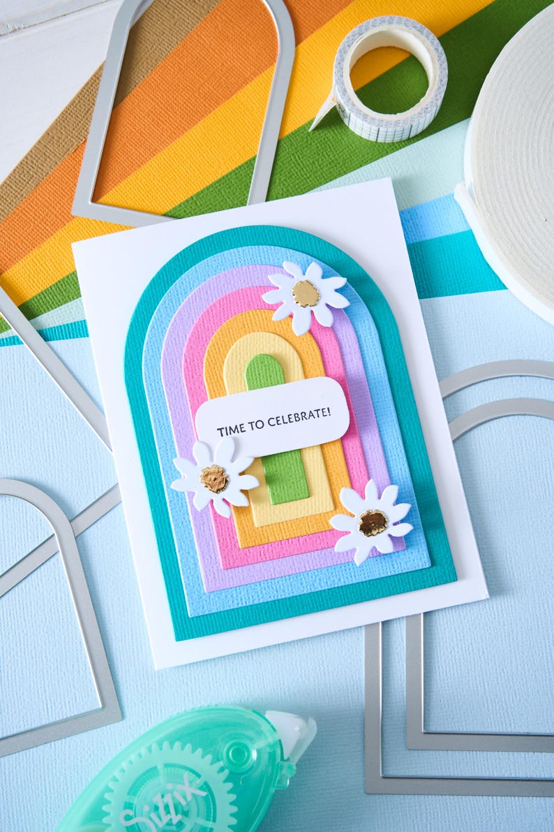 K&ouml;nnte beinhalten: Eine handgefertigte Gru&szlig;karte mit einem Regenbogenbogen-Design, mit Schichten aus farbigem Papier und wei&szlig;en G&auml;nsebl&uuml;mchen-Verzierungen. Die Karte enth&auml;lt den Text "TIME TO CELEBRATE!" und ist von Bastelmaterialien umgeben.