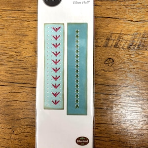 Puede incluir: Un paquete de troqueles Sizzix Thinlits Stitchlits de Eileen Hull. El paquete presenta dos troqueles rectangulares con diseños cosidos. Uno tiene flechas rojas, el otro puntadas verdes. El paquete está sobre una superficie de madera.