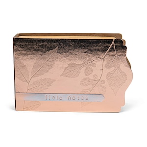May include: A rose gold notepad with a leaf design and the text "field notes" on the front.