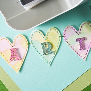 Peut inclure: Une guirlande d&eacute;corative avec des pendentifs en forme de c&oelig;ur &eacute;pelant le mot "ART". Chaque pendentif est de couleur pastel avec une bordure cousue et une lettre d&eacute;corative. La guirlande est suspendue &agrave; un fil vert, id&eacute;ale pour un projet cr&eacute;atif.