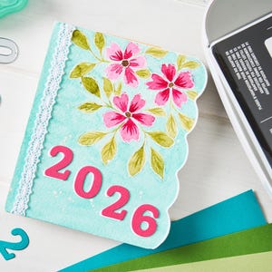 May include: A light blue, scalloped-edge journal cover with the year "2026" in pink. The cover is decorated with pink flowers and green leaves. White lace trim is along the spine. Teal and gray number cutouts are scattered around the journal.