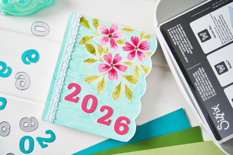 May include: A light blue, scalloped-edge journal cover with the year "2026" in pink. The cover is decorated with pink flowers and green leaves. White lace trim is along the spine. Teal and gray number cutouts are scattered around the journal.