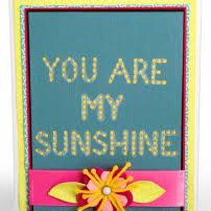 Puede incluir: Tarjeta de felicitación con fondo turquesa y el texto "YOU ARE MY SUNSHINE" en letras punteadas amarillas. Una cinta rosa y una flor decorativa con pétalos amarillos y centro blanco en la parte inferior. La tarjeta tiene un borde amarillo y verde.