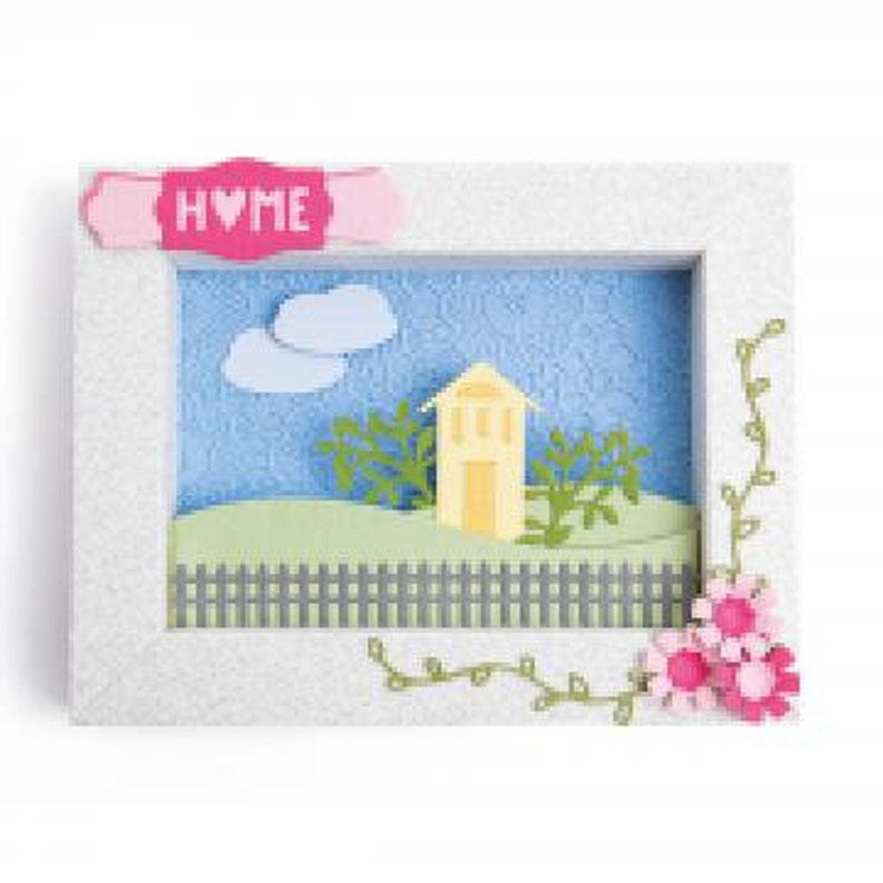 Op de afbeelding: Een wit frame met een 3D-papieren sc&egrave;ne erin. De sc&egrave;ne toont een geel huis met een groen dak, groene struiken, een grijs hek en twee witte wolken tegen een blauwe lucht. Het frame heeft een roze banner bovenaan met de tekst "HOME".