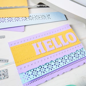 May include: A handmade greeting card with the word "HELLO" in large, light purple letters. The card features layers of yellow, light purple, and blue paper with decorative cut-out edges. A craft machine and glue are visible.